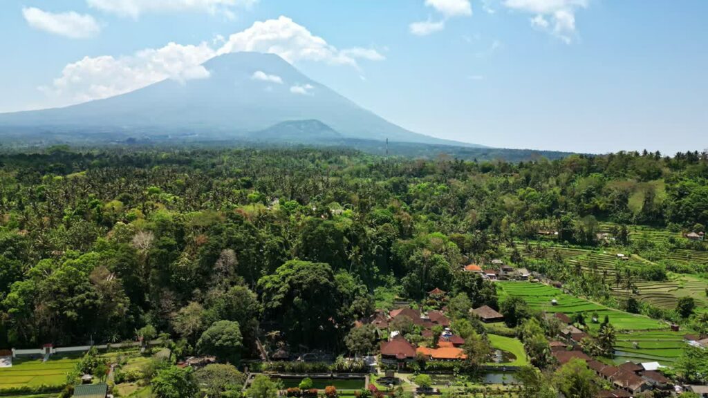 Video - Aerial drone view of Mount Agung volcano rising above palm forests and rice fields in East Bali, Indonesia. Hazy tropical atmosphere with traditional Balinese villages in the foreground