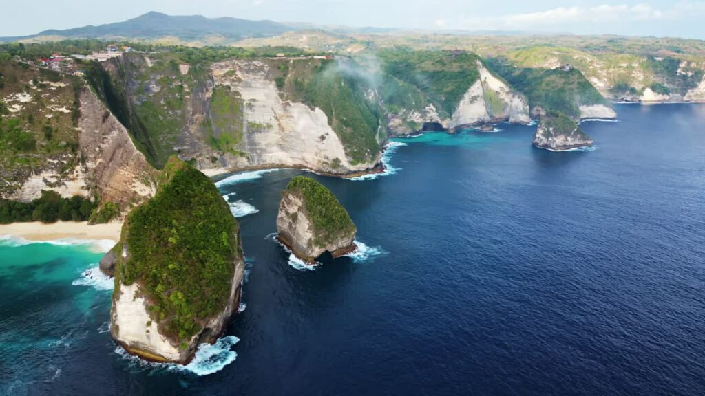 Video - Aerial drone view of Kelingking Beach in Nusa Penida, Bali. Massive limestone cliffs and offshore rock formations rise from deep blue ocean waters