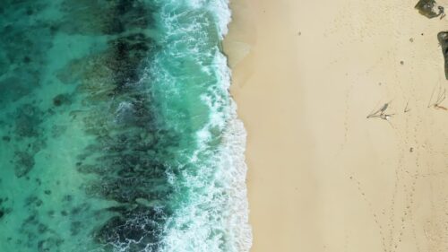 Video - Aerial drone view of turquoise ocean waves rolling onto a sandy tropical beach in Bali, Indonesia. Clear water, coral reef patterns, and soft surf create a peaceful coastal landscape