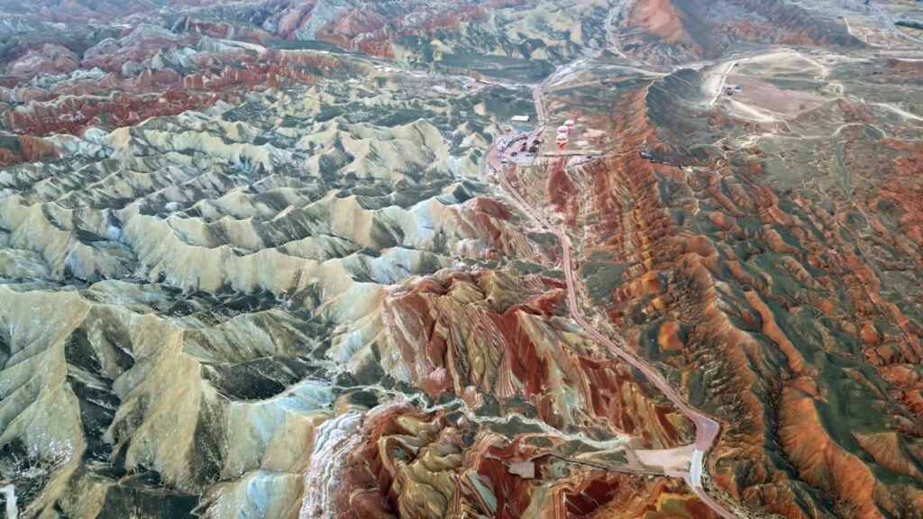 Video - Aerial drone view of Zhangye Danxia National Geopark, Zhangye, Gansu, China. Winding desert road cuts through multicolored striped hills while hot air balloons prepare for launch near the scenic viewpoint