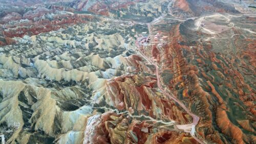 Video - Aerial drone view of Zhangye Danxia National Geopark, Zhangye, Gansu, China. Winding desert road cuts through multicolored striped hills while hot air balloons prepare for launch near the scenic viewpoint