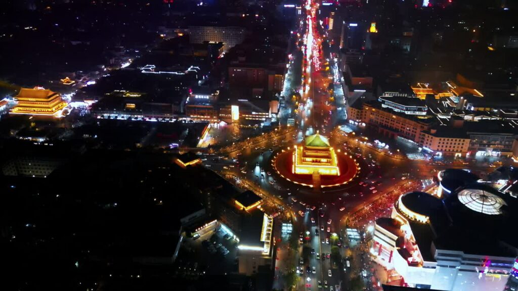 Video - Aerial drone view of the Xi'an Bell Tower brightly illuminated at night in the center of a circular intersection. Heavy traffic creates dynamic light trails around the historic Ming Dynasty structure in downtown Xi'an