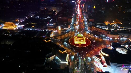Video - Aerial drone view of the Xi'an Bell Tower brightly illuminated at night in the center of a circular intersection. Heavy traffic creates dynamic light trails around the historic Ming Dynasty structure in downtown Xi'an