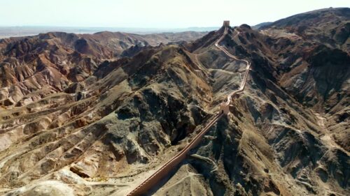 Video - Aerial drone view of the Great Wall of China winding along arid mountain ridges in Gansu Province. The ancient stone fortification stretches across barren hills under clear daylight, showcasing China's historic defensive architecture in a remote desert landscape