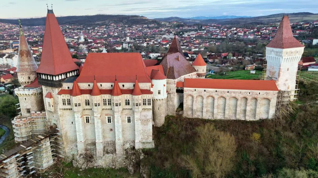 Video - Aerial drone view of Corvin Castle in Hunedoara, Transylvania. Medieval Gothic fortress with red tiled roofs, stone towers, and dramatic defensive walls rising above the town