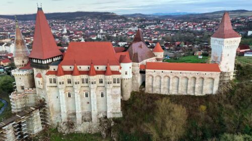 Video - Aerial drone view of Corvin Castle in Hunedoara, Transylvania. Medieval Gothic fortress with red tiled roofs, stone towers, and dramatic defensive walls rising above the town