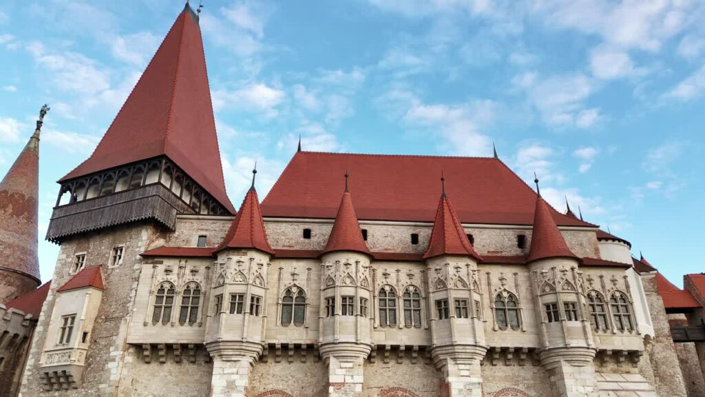 Video - Aerial drone view of Corvin Castle in Hunedoara, Transylvania. Medieval Gothic fortress with red tiled roofs, stone towers, and dramatic defensive walls rising above the town