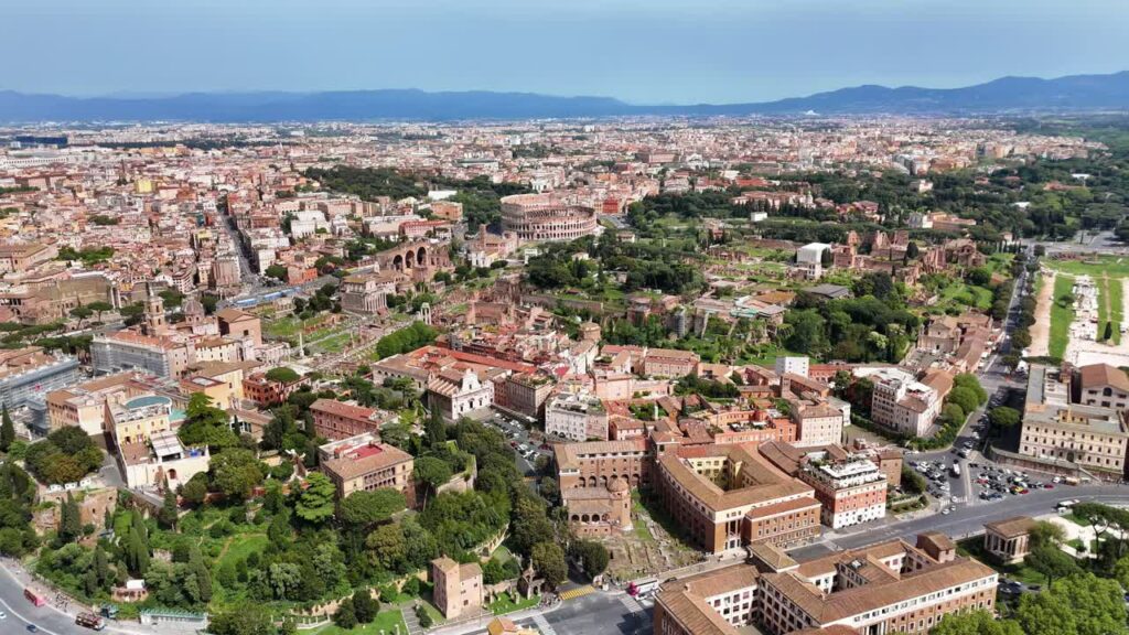 Video - Aerial drone view of Rome's historic center featuring the Colosseum, Roman Forum, and Palatine Hill. Ancient ruins surrounded by modern cityscape under clear daylight