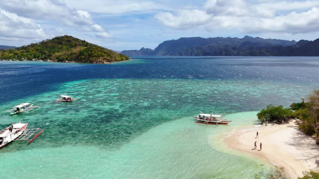 Video - Aerial drone view of traditional Filipino bangka boats anchored near a white sand beach in Coron, Palawan, Philippines. Crystal clear shallow waters transition from light aqua to deep blue with limestone islands in the distance