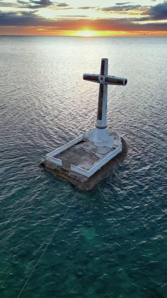 Video - Aerial drone view of the iconic Sunken Cemetery Cross monument off the coast of Camiguin Island, Philippines. Large white cross stands on a submerged platform in the ocean at sunset, marking a historic volcanic eruption site in northern Mindanao. Vertical