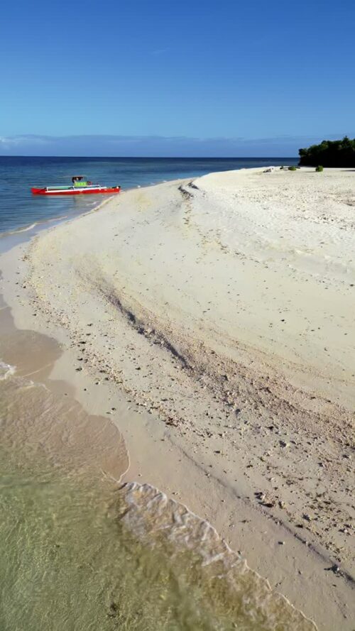 Video - Aerial drone view of a pristine white sand beach in northern Palawan, Philippines, with calm turquoise water and a small orange boat resting near shore. Vertical