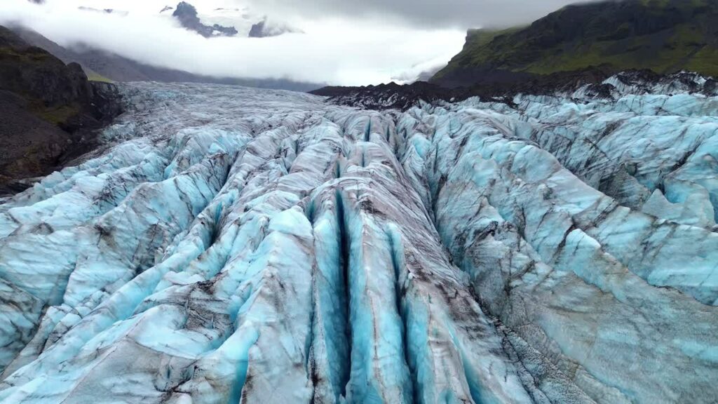 Video - Aerial drone view of deep crevasses of Falljokull glacier, an outlet of Vatnajokull in South Iceland. Dramatic blue ice ridges and dark volcanic ash streaks create powerful Arctic landscape scenery under cloudy sky