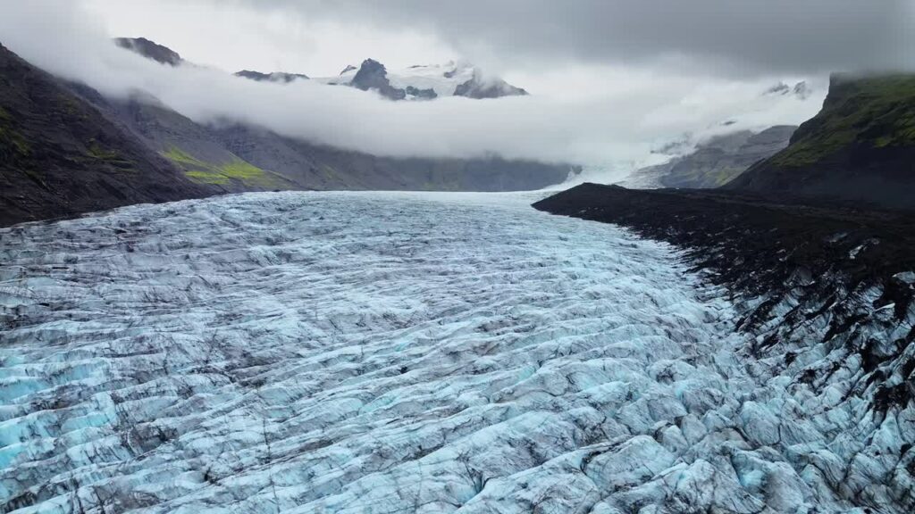 Video - Aerial drone view of Vatnajokull glacier flowing through a rugged valley in Skaftafell, South Iceland. Fog rolls over mountain peaks while the expansive ice sheet stretches toward the horizon in dramatic Nordic scenery