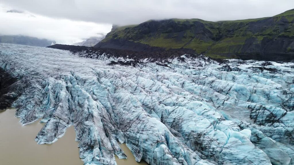 Video - Aerial drone view of deep crevasses of Falljokull glacier, an outlet of Vatnajokull in South Iceland. Dramatic blue ice ridges and dark volcanic ash streaks create powerful Arctic landscape scenery under cloudy sky