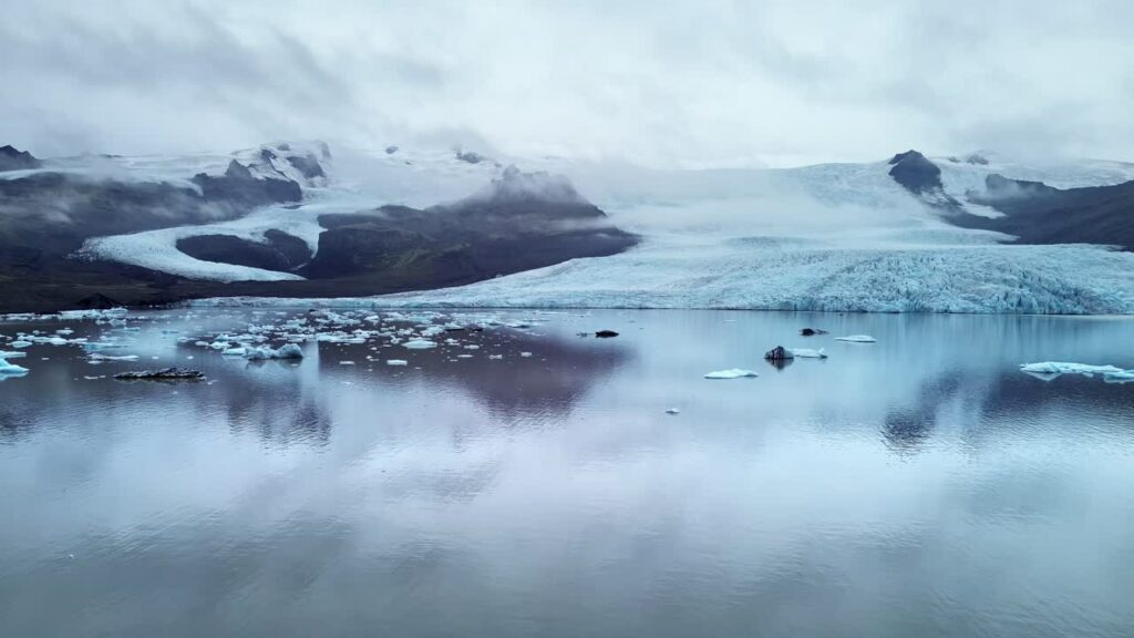 Video - Aerial drone view of Jokulsarlon Glacier Lagoon in Southeast Iceland. Floating icebergs drift across calm reflective waters beneath Vatnajokull ice cap