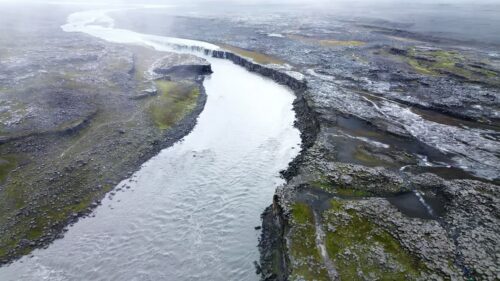 Video - Aerial drone view of the Jokulsa a Fjollum river winding through a barren volcanic plateau near Dettifoss. Moss covered rocks and dark lava formations frame the powerful glacial river