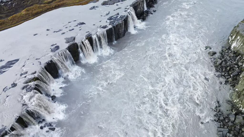 Video - Aerial drone view of Dettifoss waterfall cascading over dark volcanic cliffs. Heavy glacial currents crash into the canyon below