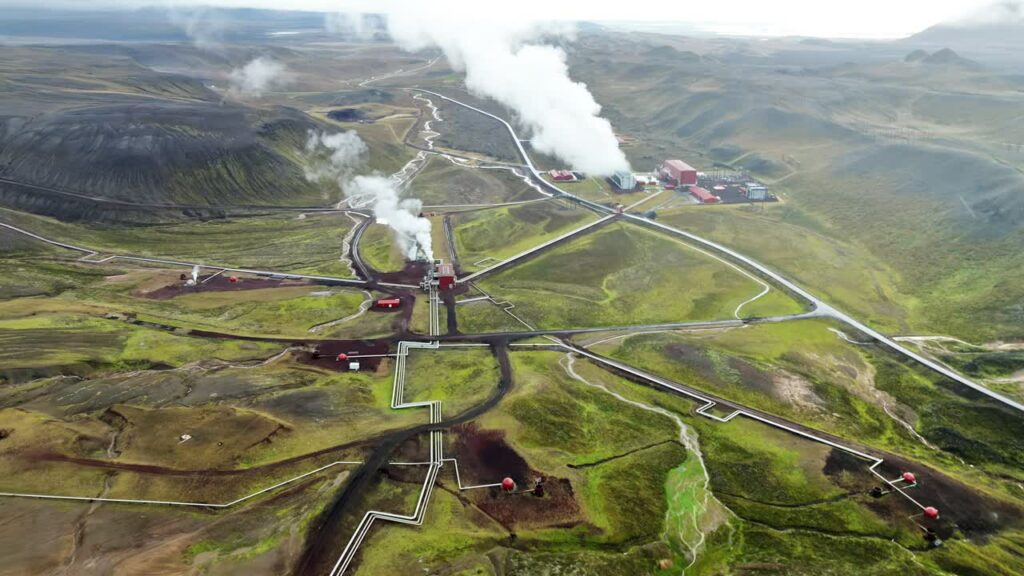 Video - Aerial drone view of Krafla geothermal power station with steam rising from pipelines across rolling volcanic hills. Renewable energy infrastructure set within Iceland's dramatic geothermal landscape