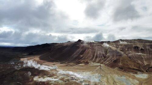 Video - Aerial drone view of steaming fumaroles and sulfur vents in the Hverir geothermal area near Namafjall. White steam rises from mineral stained earth in an otherworldly volcanic environment