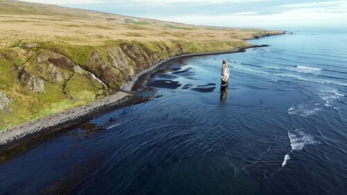 Video - Aerial drone view of Hvitserkur, the famous basalt sea stack rising from the ocean along the Vatnsnes Peninsula in northern Iceland. Waves gently roll across the black sand beach while cliffs and grassy coastal plains stretch along the shoreline