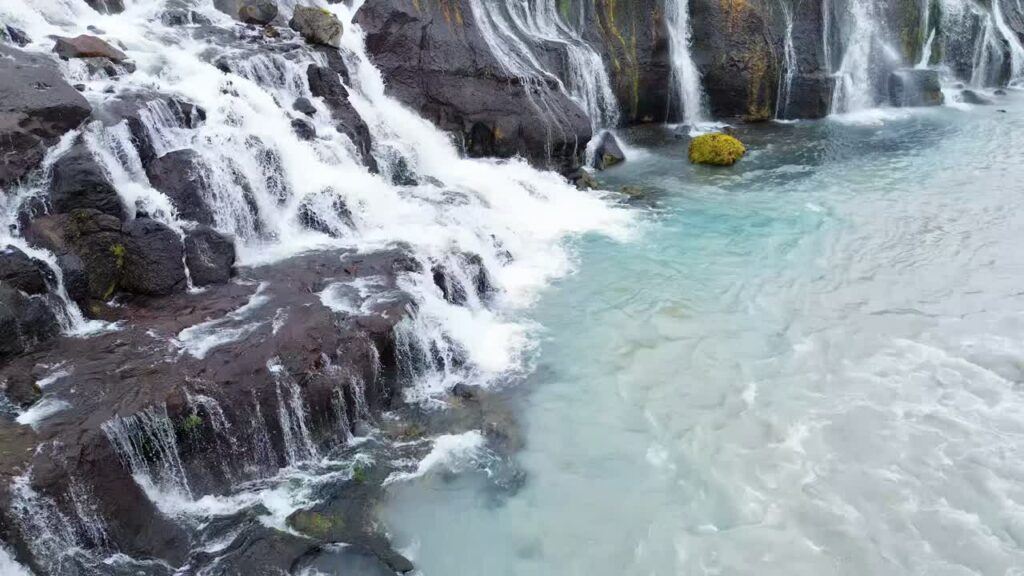 Video - Aerial drone view of rocky ledges of Hraunfossar, capturing water flowing through volcanic lava formations before dropping into the turquoise river below
