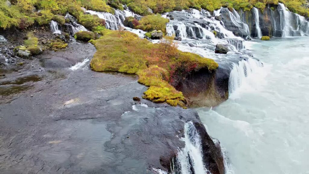 Video - Aerial drone view of rocky ledges of Hraunfossar, capturing water flowing through volcanic lava formations before dropping into the turquoise river below