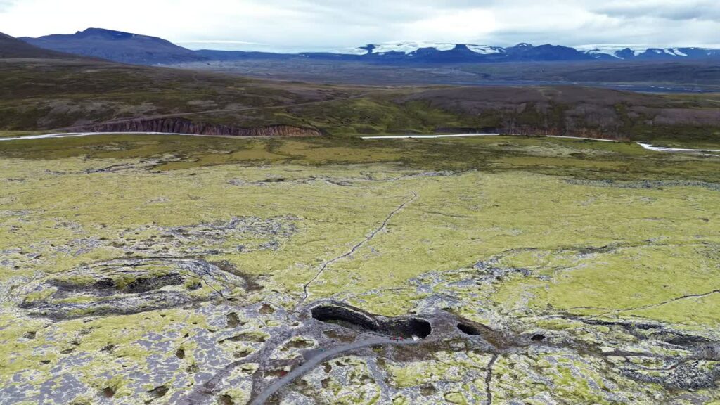 Video - Aerial drone view an expansive moss covered lava field with volcanic caves and openings scattered across the terrain. Distant mountains and glaciers are visible on the horizon of the Icelandic highlands