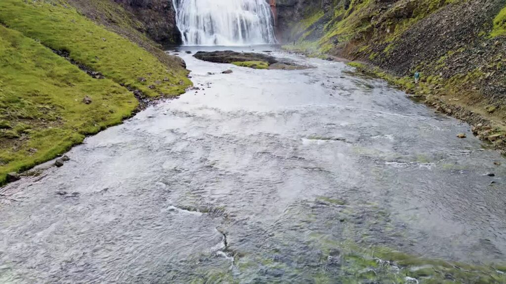 Video - Aerial drone view of a large waterfall at the end of a river channel surrounded by moss covered cliffs and rocky slopes in Iceland's remote landscape