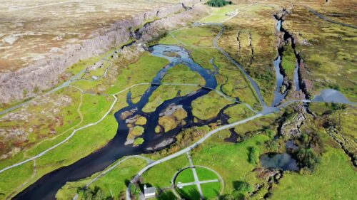 Video - Aerial drone view of the Pingvellir National Park rift valley where tectonic plates separate. Rivers and wetlands weave through green plains surrounded by dramatic rock cliffs and walking paths