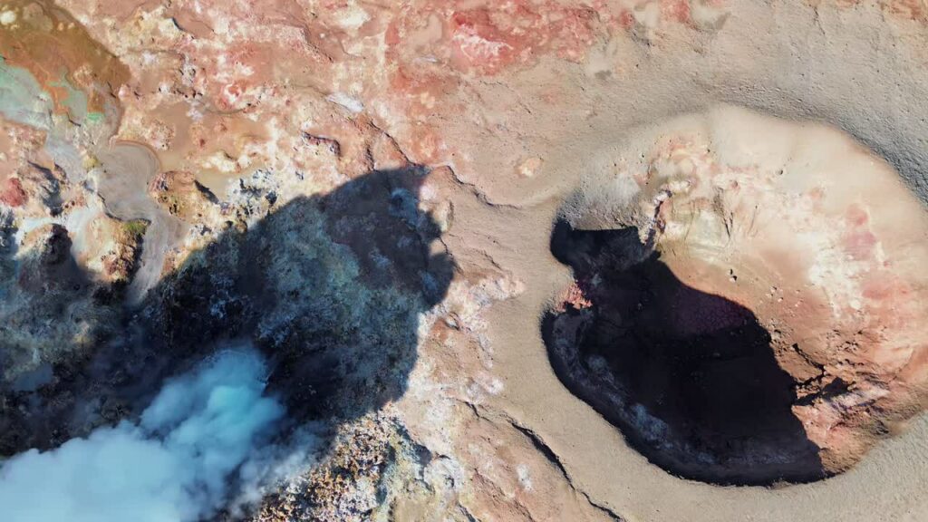 Video - Aerial drone view of steaming geothermal vents and bubbling mud pools in a volcanic geothermal field in Iceland. Mineral rich ground in shades of beige, red, and blue surrounds active fumaroles releasing steam