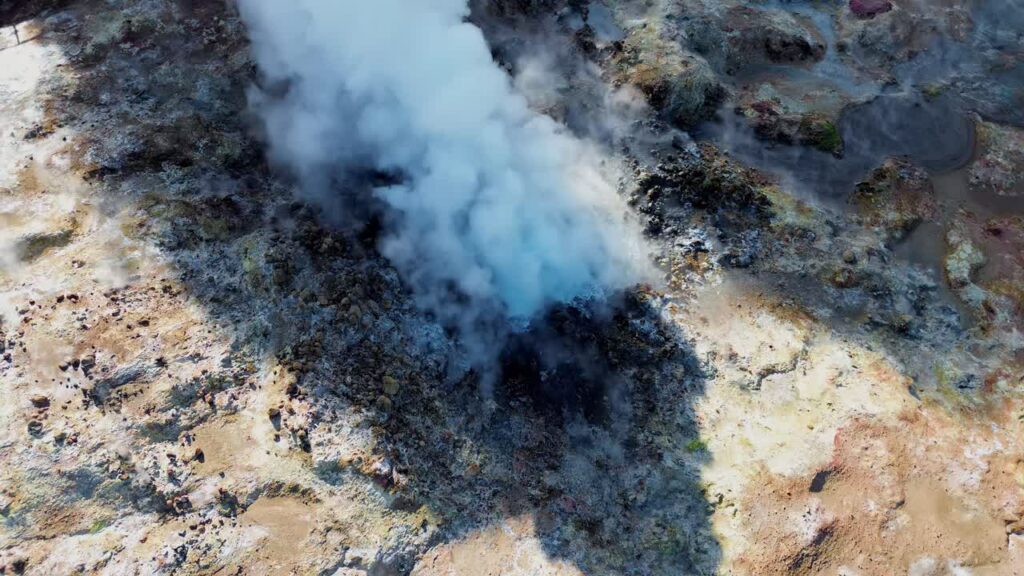 Video - Aerial drone view of steaming geothermal vents and bubbling mud pools in a volcanic geothermal field in Iceland. Mineral rich ground in shades of beige, red, and blue surrounds active fumaroles releasing steam