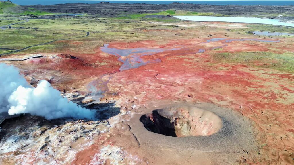 Video - Aerial drone view of steaming geothermal vents and bubbling mud pools in a volcanic geothermal field in Iceland. Mineral rich ground in shades of beige, red, and blue surrounds active fumaroles releasing steam