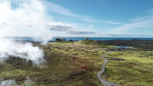 Video - Aerial drone view of steaming geothermal vents across the rugged lava fields of the Reykjanes Peninsula. A winding road leads toward a lighthouse near the Atlantic Ocean under clear blue skies
