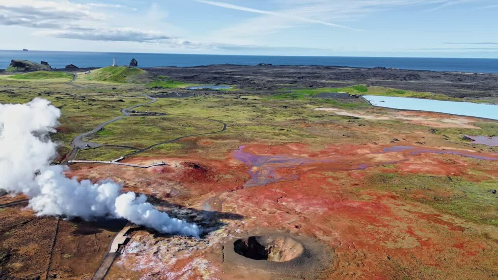 Video - Aerial drone view of steaming geothermal vents and bubbling mud pools in a volcanic geothermal field in Iceland. Mineral rich ground in shades of beige, red, and blue surrounds active fumaroles releasing steam