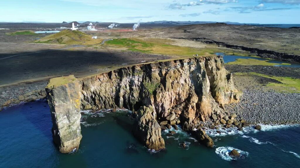 Video - Aerial drone view of moss covered coastal cliffs and a rocky beach along Iceland's volcanic shoreline. Turquoise ocean waves break against the steep basalt cliffs