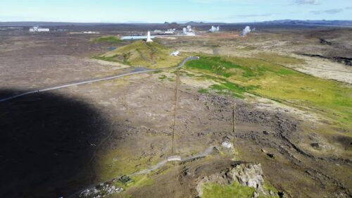 Video - Aerial drone view of Reykjanes lighthouse standing on a grassy hill surrounded by vast volcanic lava fields. Steam plumes from nearby geothermal areas rise in the distance