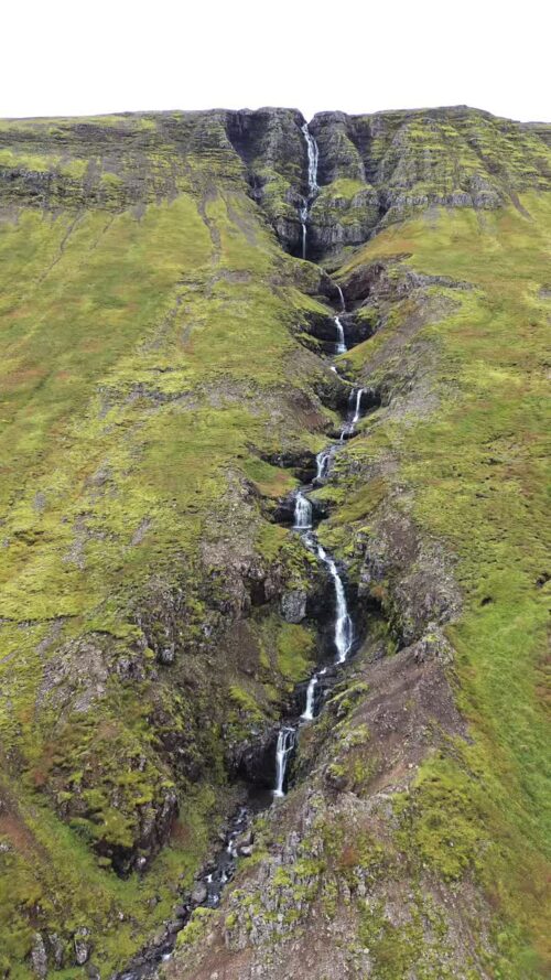 Video - Aerial drone view of a long cascading waterfall flowing down a steep green mountainside in Iceland. Multiple tiers of water descend through moss covered volcanic rock. Vertical
