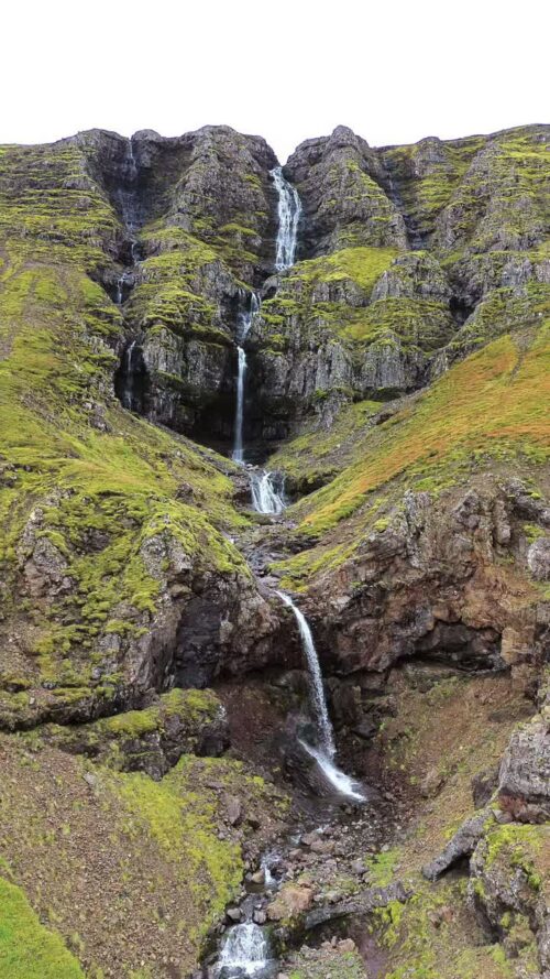 Video - Aerial drone view of a long cascading waterfall flowing down a steep green mountainside in Iceland. Multiple tiers of water descend through moss covered volcanic rock. Vertical