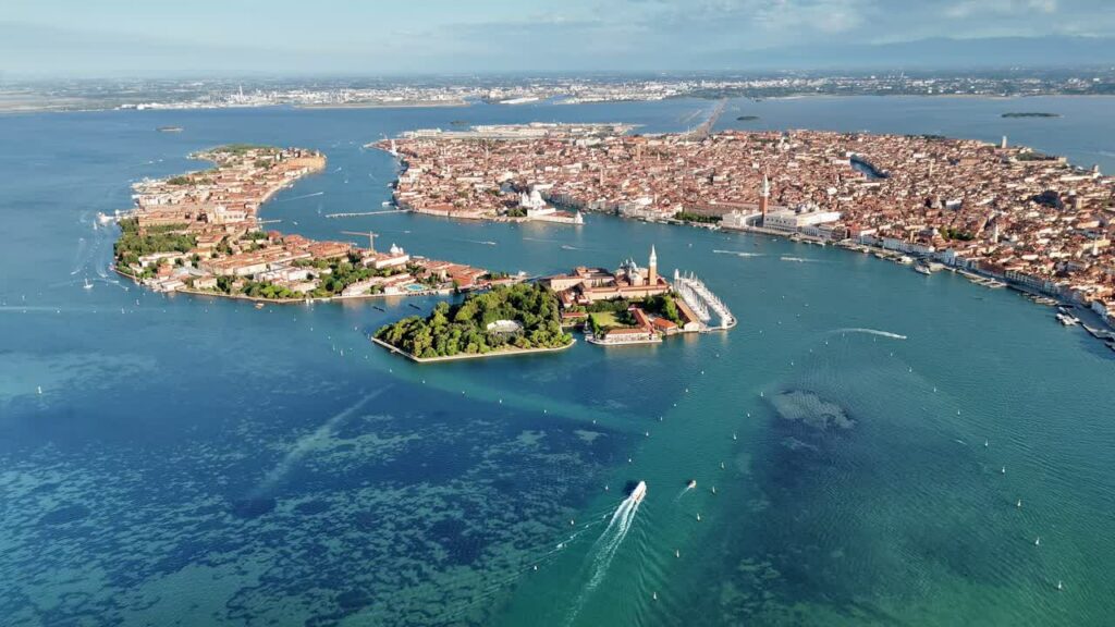Video - Aerial drone view of the Venice Lagoon with San Giorgio Maggiore island and St. Mark's Basin. Boats move through turquoise water while the historic skyline of Venice spreads across the horizon