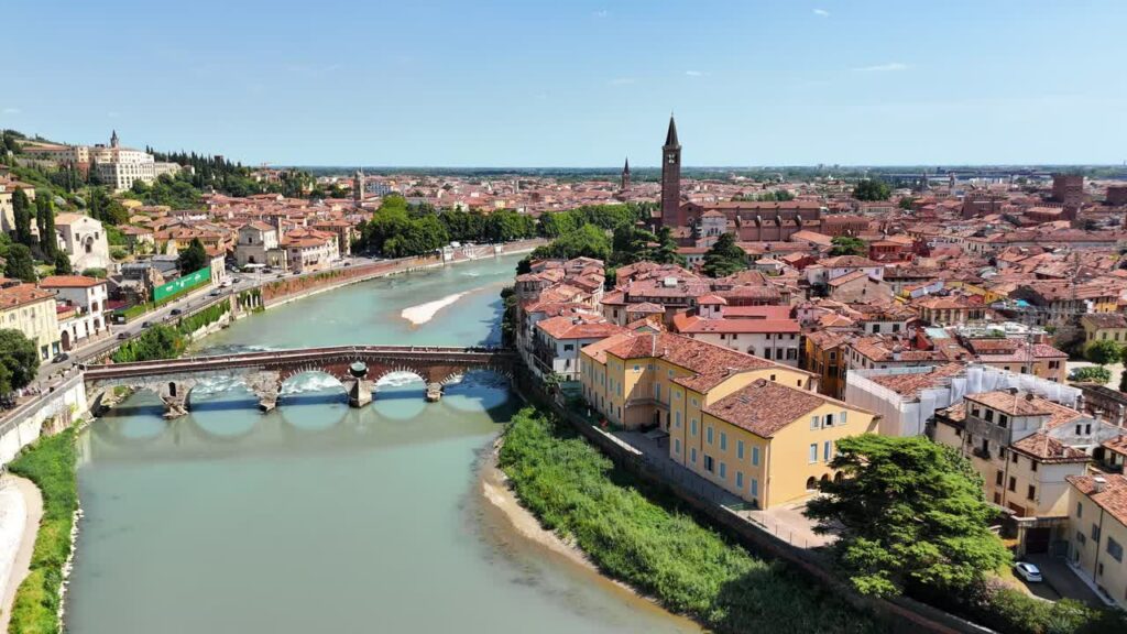 Video - Aerial drone view of Verona's historic center with the Adige River winding through the city. A stone bridge crosses the river near terracotta rooftops and medieval towers