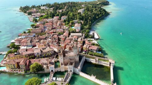 Video - Aerial drone view of Scaliger Castle in Sirmione surrounded by turquoise waters of Lake Garda. Medieval walls and towers rise above the harbor and historic lakeside town