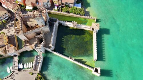Video - Aerial drone view of Scaliger Castle in Sirmione with its fortified walls enclosing a small harbor on the crystal clear waters of Lake Garda