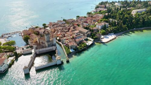 Video - Aerial drone view of Scaliger Castle in Sirmione surrounded by turquoise waters of Lake Garda. Medieval walls and towers rise above the harbor and historic lakeside town