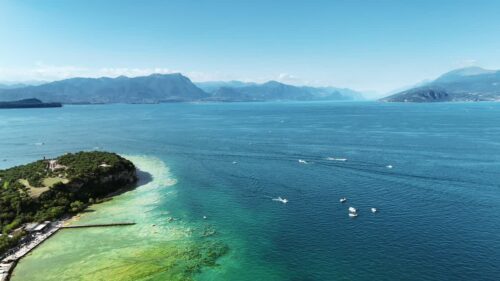 Video - Aerial drone view the turquoise waters of Lake Garda with a small island and boats moving across the lake. The dramatic alpine mountains rise in the background under a clear blue sky in northern Italy