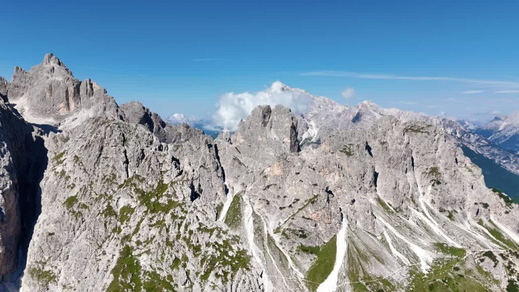 Video - Aerial drone view of the dramatic Dolomites mountain range with steep grey cliffs and alpine valleys below. Clouds drift over the rugged peaks of northern Italy