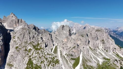 Video - Aerial drone view of the dramatic Dolomites mountain range with steep grey cliffs and alpine valleys below. Clouds drift over the rugged peaks of northern Italy