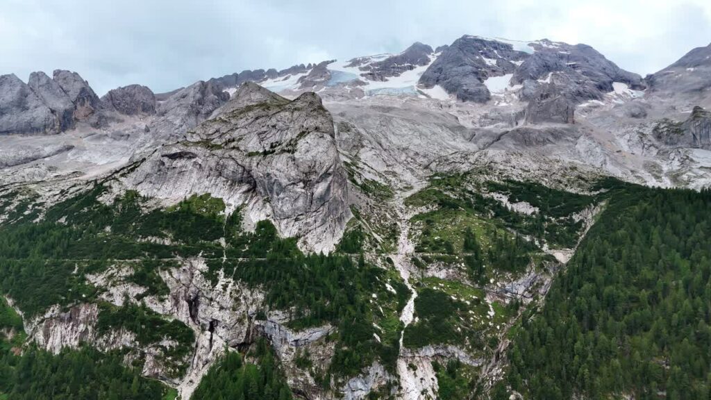 Video - Aerial drone view of rugged alpine mountains with patches of snow and glacier ice on the upper slopes. Forested valleys stretch below the massive limestone cliffs