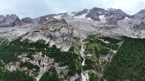 Video - Aerial drone view of rugged alpine mountains with patches of snow and glacier ice on the upper slopes. Forested valleys stretch below the massive limestone cliffs