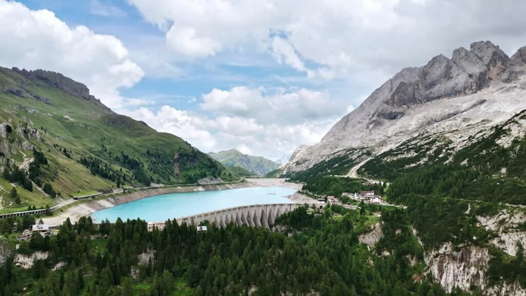Video - Aerial drone view of a curved concrete dam holding back a turquoise alpine reservoir surrounded by forested mountains and dramatic cliffs in the Italian Alps