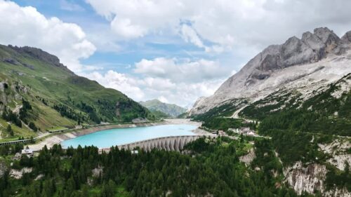 Video - Aerial drone view of a curved concrete dam holding back a turquoise alpine reservoir surrounded by forested mountains and dramatic cliffs in the Italian Alps